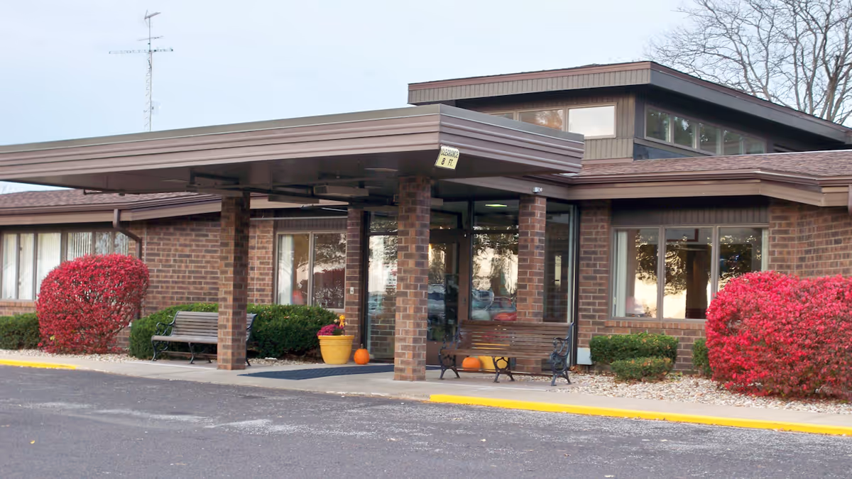 Entrance of a brick senior living building with a covered drop-off canopy, benches, potted plants and red shrubs.
