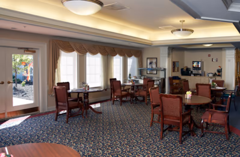 A dining room with multiple round tables and red upholstered chairs. Large windows with beige curtains allow natural light to fill the room. There is a door leading outside on the left side and a beverage station with coffee machines and supplies in the back corner.