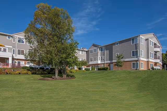 Exterior view of a multi-story assisted living facility building with gray and white siding, surrounded by a well-maintained green lawn and a large tree in the foreground under a blue sky with some clouds.