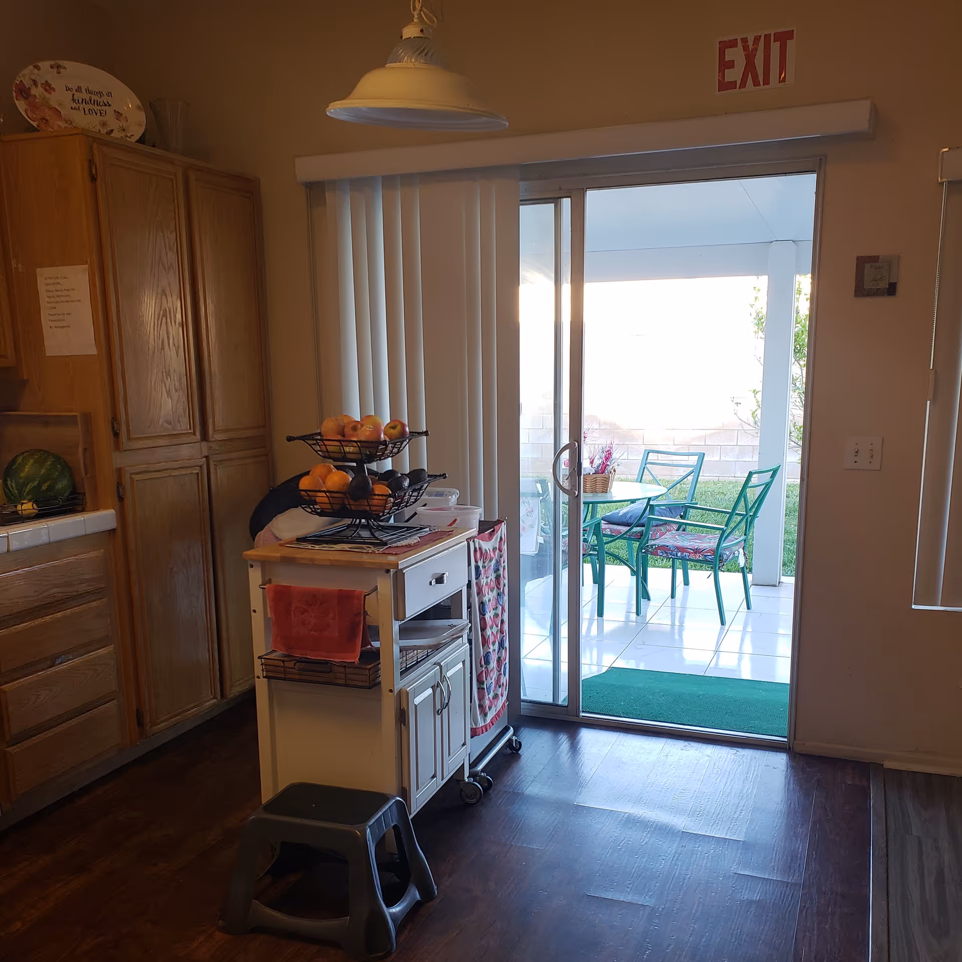 Interior view of a kitchen area with wooden cabinets on the left, a small white kitchen cart with fruit baskets and towels in the center, and a sliding glass door leading to an outdoor patio with green chairs and a table. An exit sign is visible above the door.