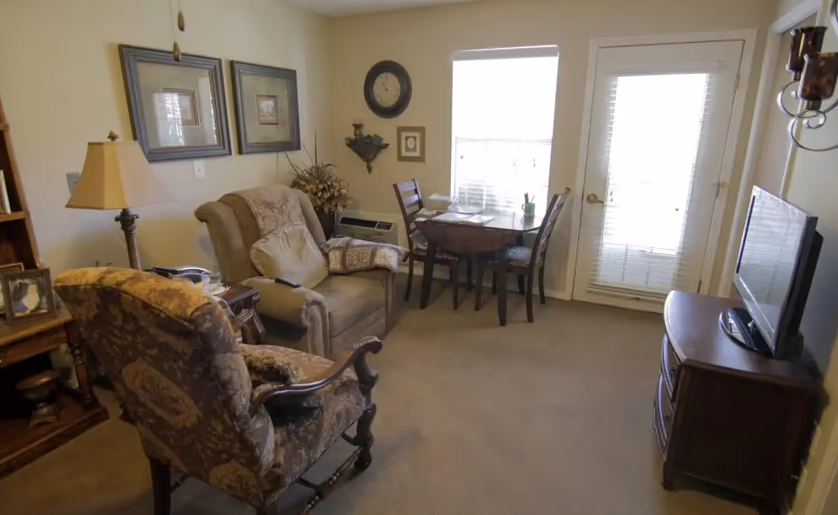 A cozy living room with two armchairs, a small wooden dining table with two chairs near a window, a TV on a wooden stand, and wall decorations including two framed pictures and a clock.