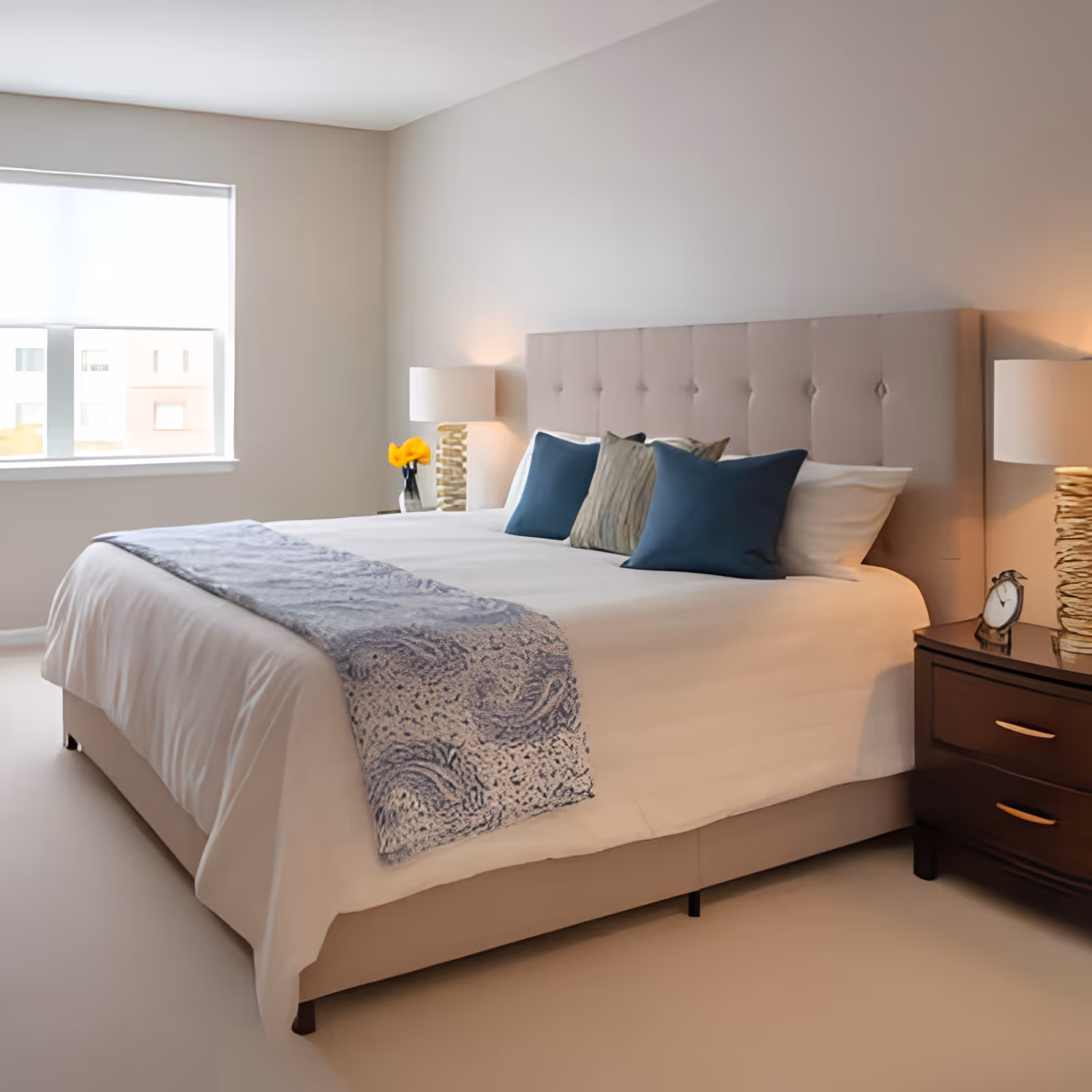A neatly made bed with a beige tufted headboard, white bedding, a blue patterned throw blanket, and several pillows in blue and beige. On each side of the bed are wooden nightstands with lamps and decorative items. A window with a white blind lets in natural light, illuminating the neutral-toned bedroom.