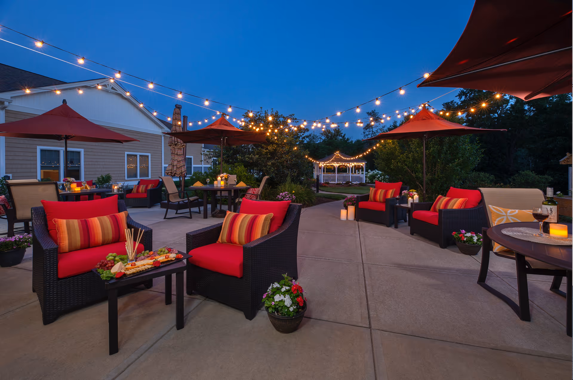 Well-lit outdoor courtyard patio at dusk with red-cushioned wicker chairs, umbrellas, string lights, and a gazebo.