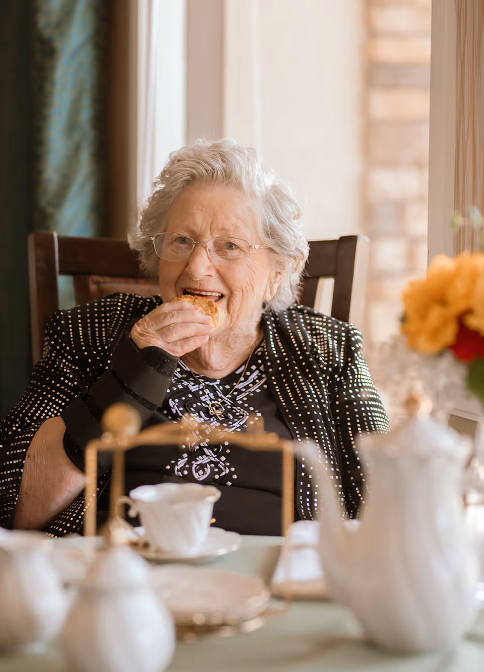Elderly woman smiling while eating a pastry at a table set with a teapot and teacup.