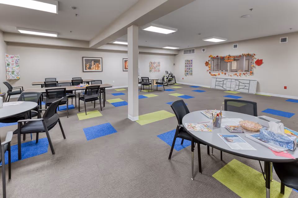 A spacious activity room in a senior living facility with multiple tables and chairs arranged around the room. The floor has a patterned carpet with blue and green squares. On the walls, there are framed pictures, colorful artwork, and a bulletin board decorated with autumn leaves and a welcome sign. The room is well-lit with ceiling lights and has a clean, organized appearance.