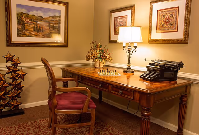 A cozy interior room featuring a wooden desk with a vintage typewriter, a chessboard, and a decorative lamp. A wooden chair with a red cushion is placed in front of the desk. The walls are adorned with framed artwork, and a decorative star-shaped sculpture stands on the floor to the left.