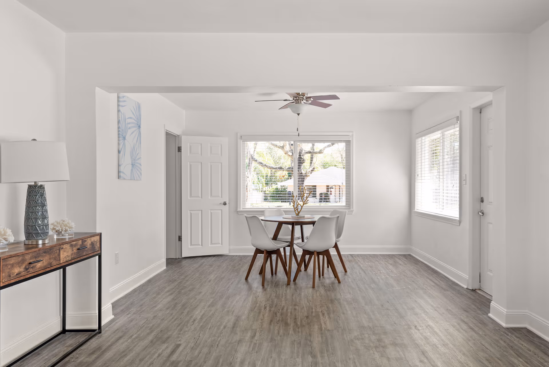 A bright dining area with a round wooden table and four white chairs with wooden legs. The room has light gray wood flooring, white walls, and large windows with white blinds letting in natural light. A ceiling fan with wooden blades is mounted above the table. To the left, there is a wooden console table with a blue patterned lamp and decorative coral pieces. A door is open on the left side of the room, and another door is visible on the right side.