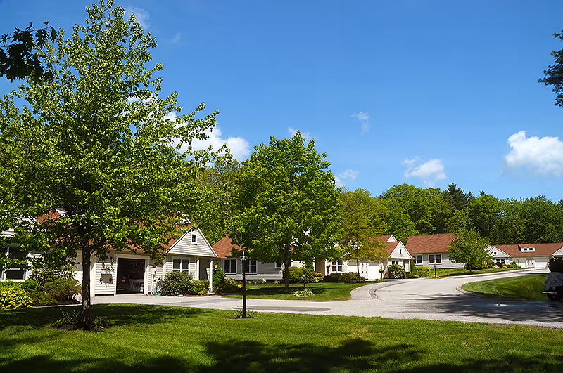 A sunny outdoor scene at a senior living facility with several single-story buildings with white siding and red roofs, surrounded by green trees and well-maintained lawns under a clear blue sky.