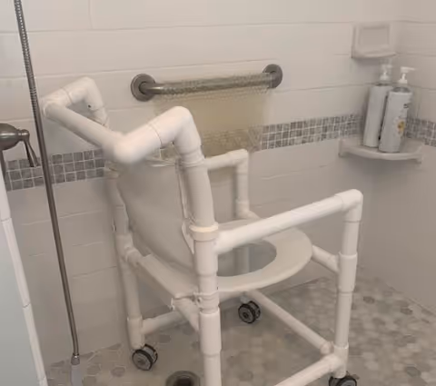 A white shower chair with wheels placed inside a tiled shower area. The shower walls have white tiles with a horizontal strip of small gray mosaic tiles. There is a grab bar on the wall and two bottles of soap or shampoo on a small wall-mounted shelf.