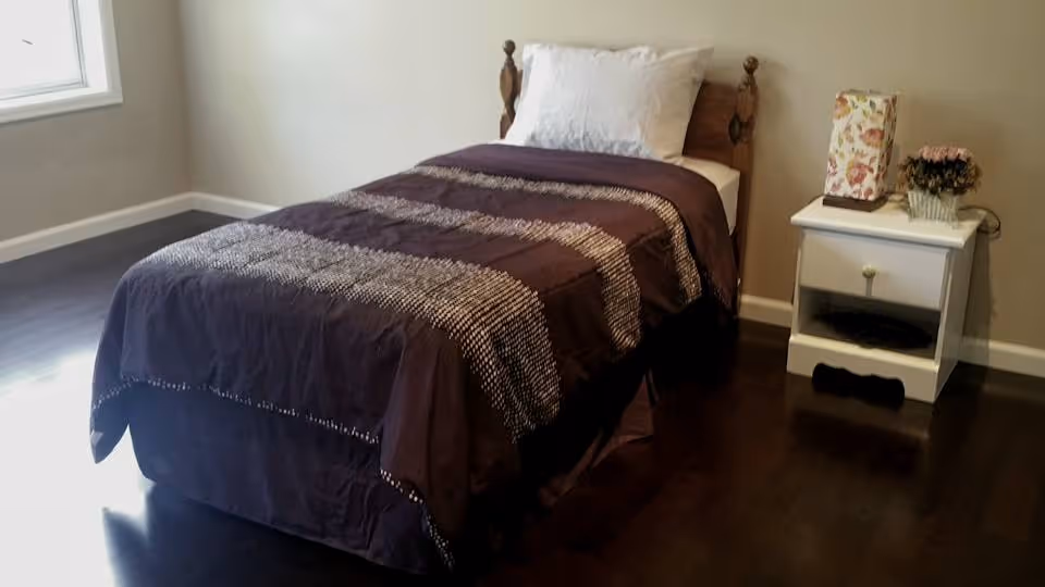A simple bedroom with a single bed covered with a dark purple blanket with white patterns, a white pillow, and a wooden headboard. Next to the bed is a white nightstand with a floral lamp and a small flower pot. The room has dark wooden flooring and a window on the left side letting in natural light.