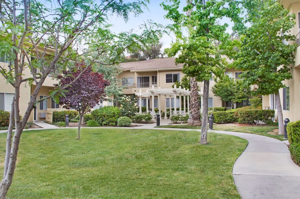 Outdoor courtyard area of a senior living facility with a well-maintained lawn, various trees including a palm tree, shrubs, and a winding concrete pathway. Beige two-story buildings with balconies and windows surround the courtyard, and a white pergola structure is visible in the center background.