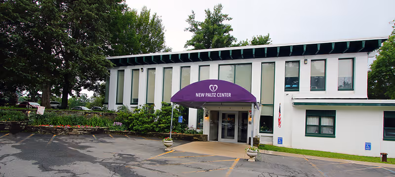 Front entrance of the New Paltz Center, a two-story white building with a purple awning, accessible parking and surrounding trees.