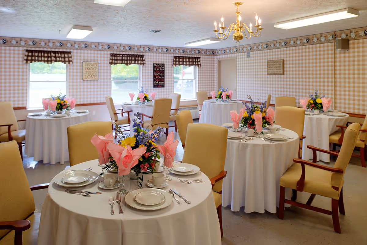 Dining room with round tables set for a meal, white tablecloths, floral centerpieces and beige chairs.