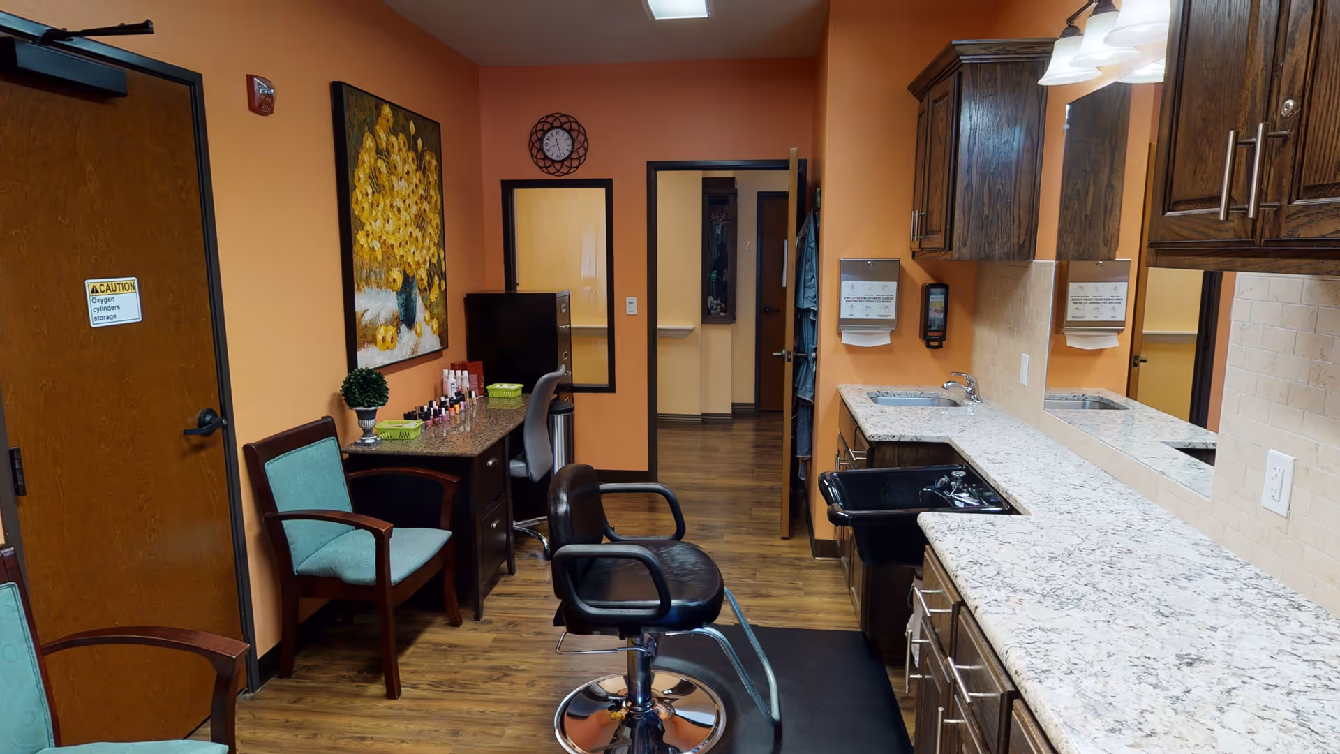 Interior view of a room with orange walls and wood flooring, featuring a black salon chair in front of a long granite countertop with a sink. There are wooden cabinets above the countertop, a desk with a chair and nail polish bottles, a green upholstered chair, a wall clock, and a painting of yellow flowers on the wall.