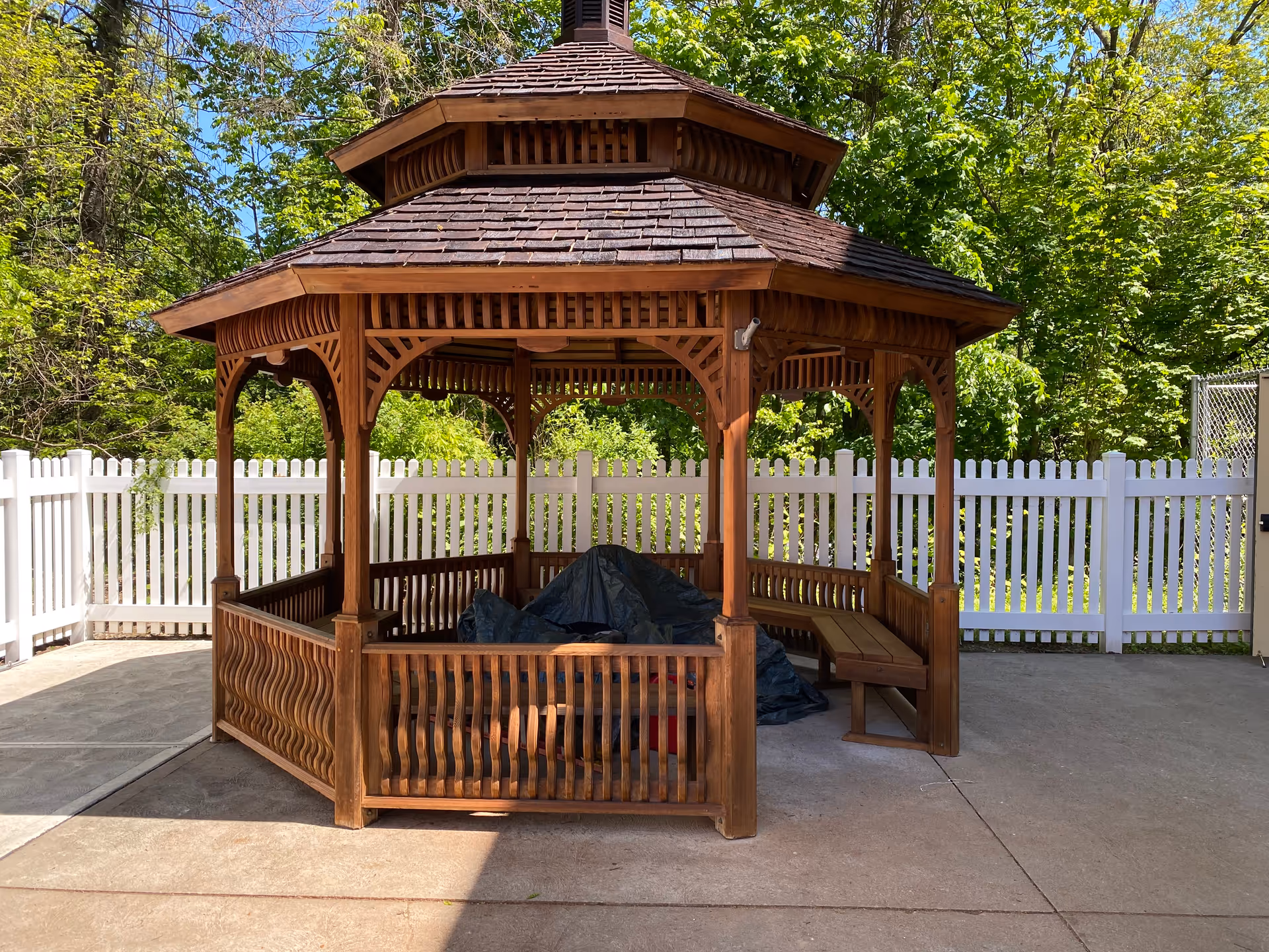 A wooden gazebo with a shingled roof and built-in benches inside a fenced outdoor area with green trees in the background. There is a tarp covering some items inside the gazebo.