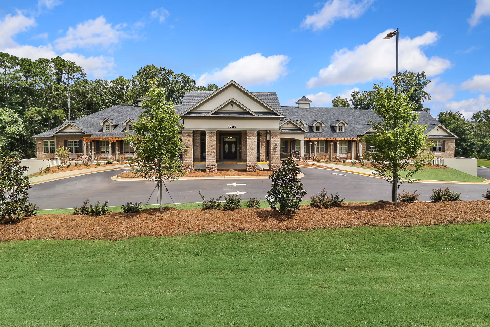 Front exterior view of a single-story senior living facility building with a driveway, landscaped greenery, and trees under a partly cloudy blue sky.