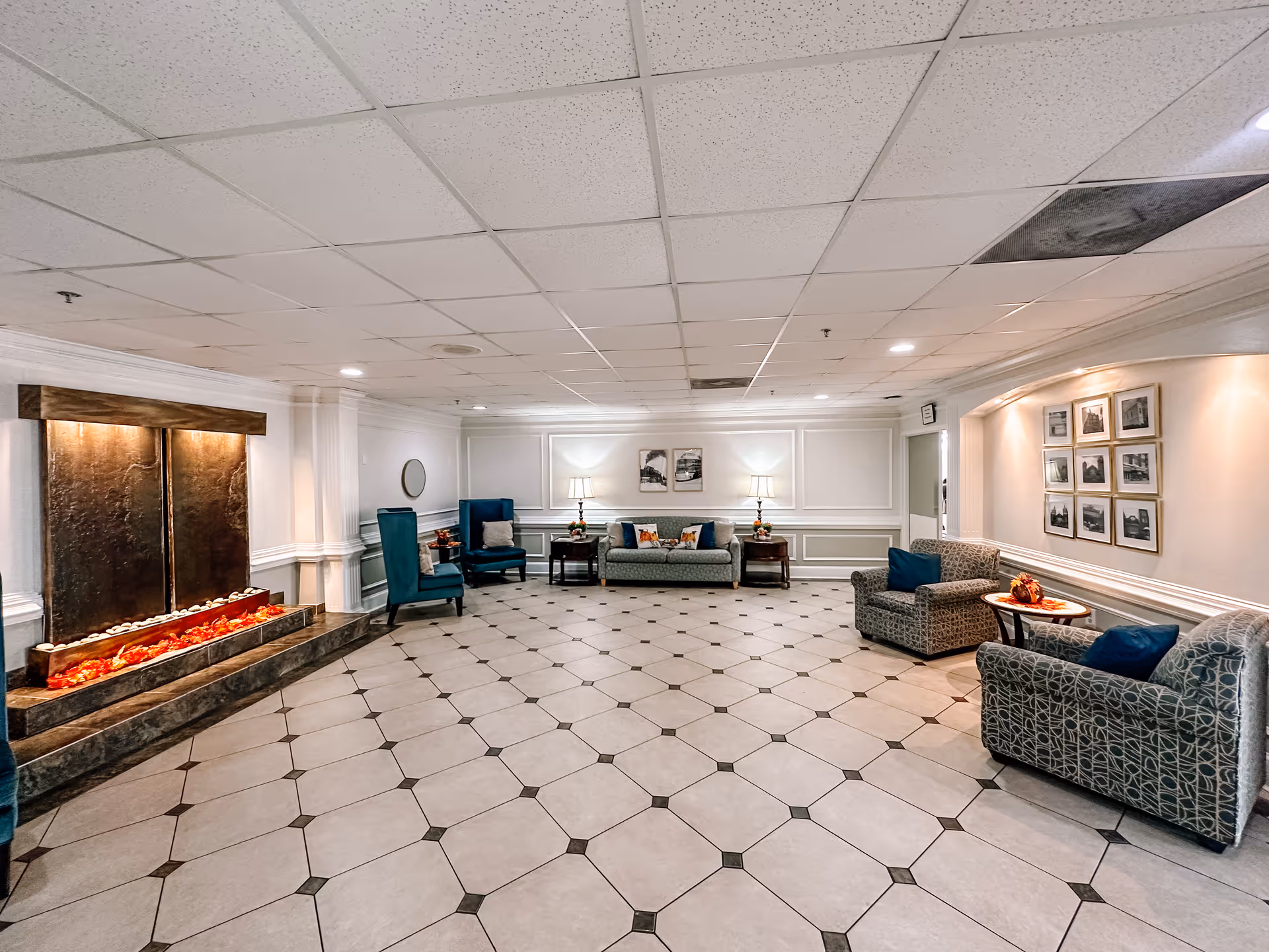 Spacious furnished lounge with tiled floor, seating areas, lamps and a decorative indoor waterfall/fireplace.