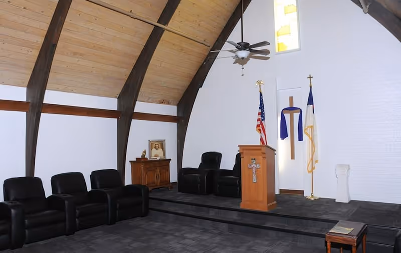 Small chapel-style interior with curved wooden beams, a pulpit, flags, and rows of black reclining chairs.