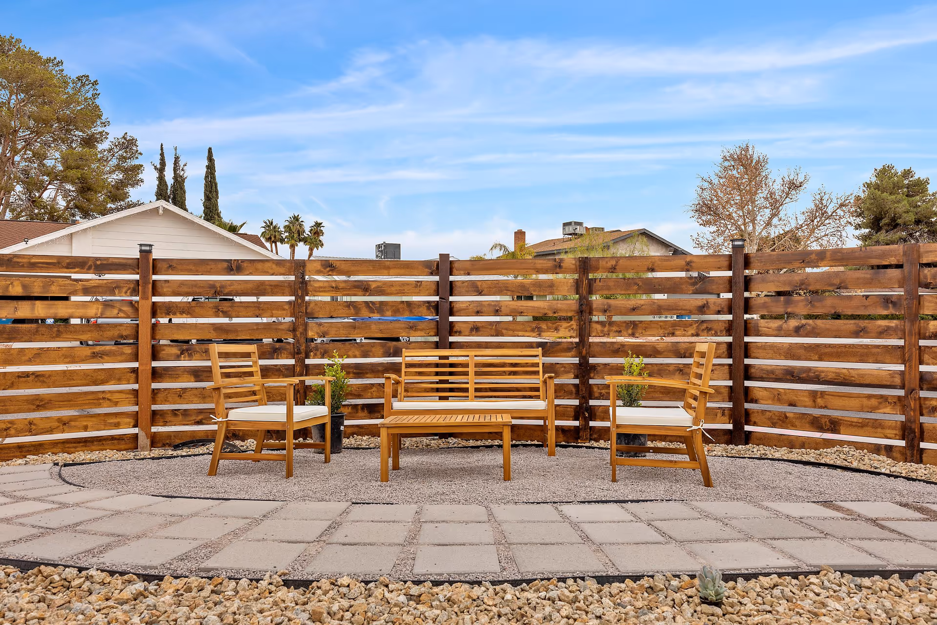 A small outdoor patio with a wooden bench, chairs and table arranged on a paved area in front of a horizontal wood-slat fence under a blue sky.