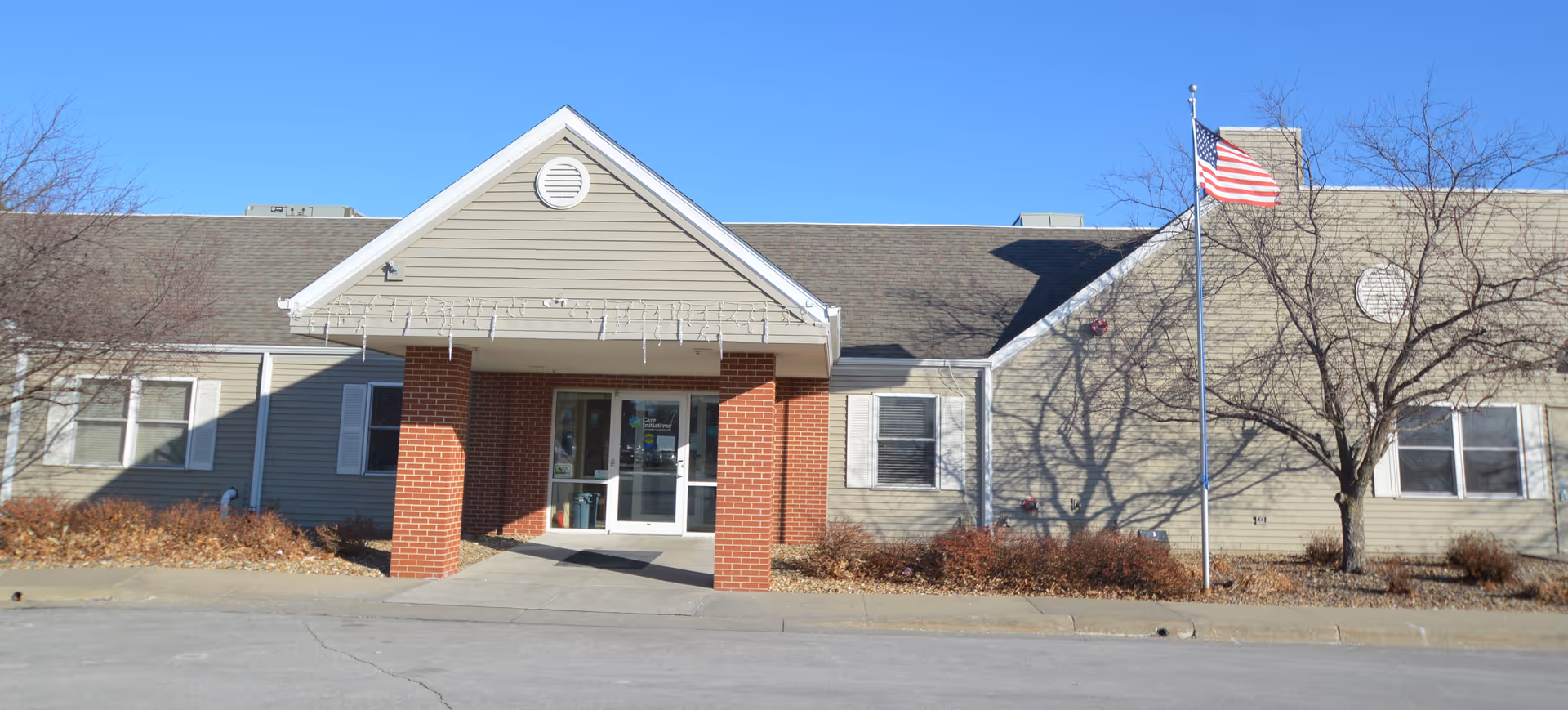 Front exterior view of Southridge Specialty Care building with a covered entrance supported by two brick columns, an American flag on a flagpole to the right, leafless trees, and clear blue sky.