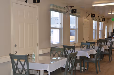 Interior view of a dining area with multiple tables covered with white tablecloths and green wooden chairs. The room has several windows with light green valances, and small black signs hanging above each table indicating table numbers. The floor is wooden, and there is a white door on the left side of the image.
