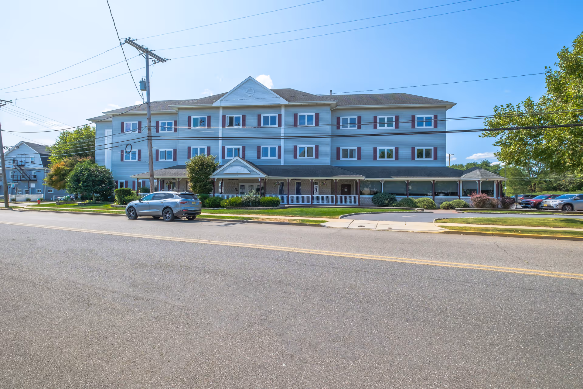 Exterior front view of a three-story senior living facility building named Bayside Manor with multiple windows, a covered porch entrance, and a parking area with cars. The building is light blue with white trim and red shutters, set against a clear blue sky.