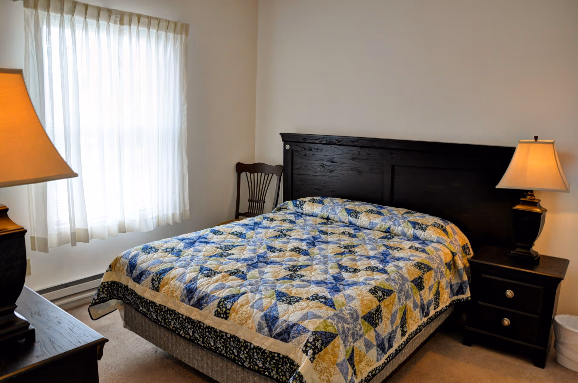 A neatly made bedroom with a patterned quilted bed, dark wood headboard and nightstands, two lamps, a window with sheer curtains, and a wooden chair.