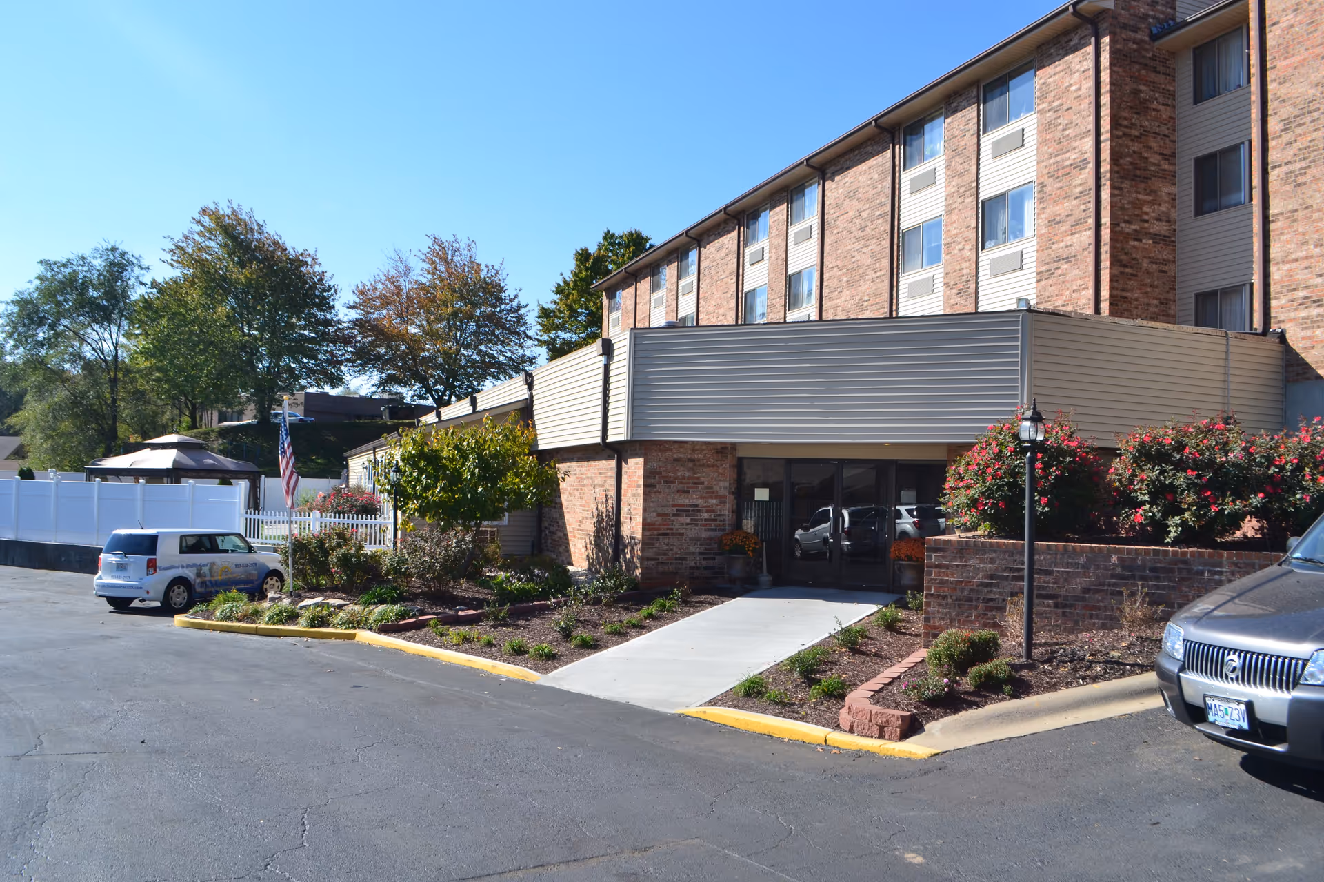 Exterior view of a multi-story senior living facility building with brick and siding facade, a paved driveway, landscaped garden beds with shrubs and flowers, an American flag on a pole, and parked cars under a clear blue sky.