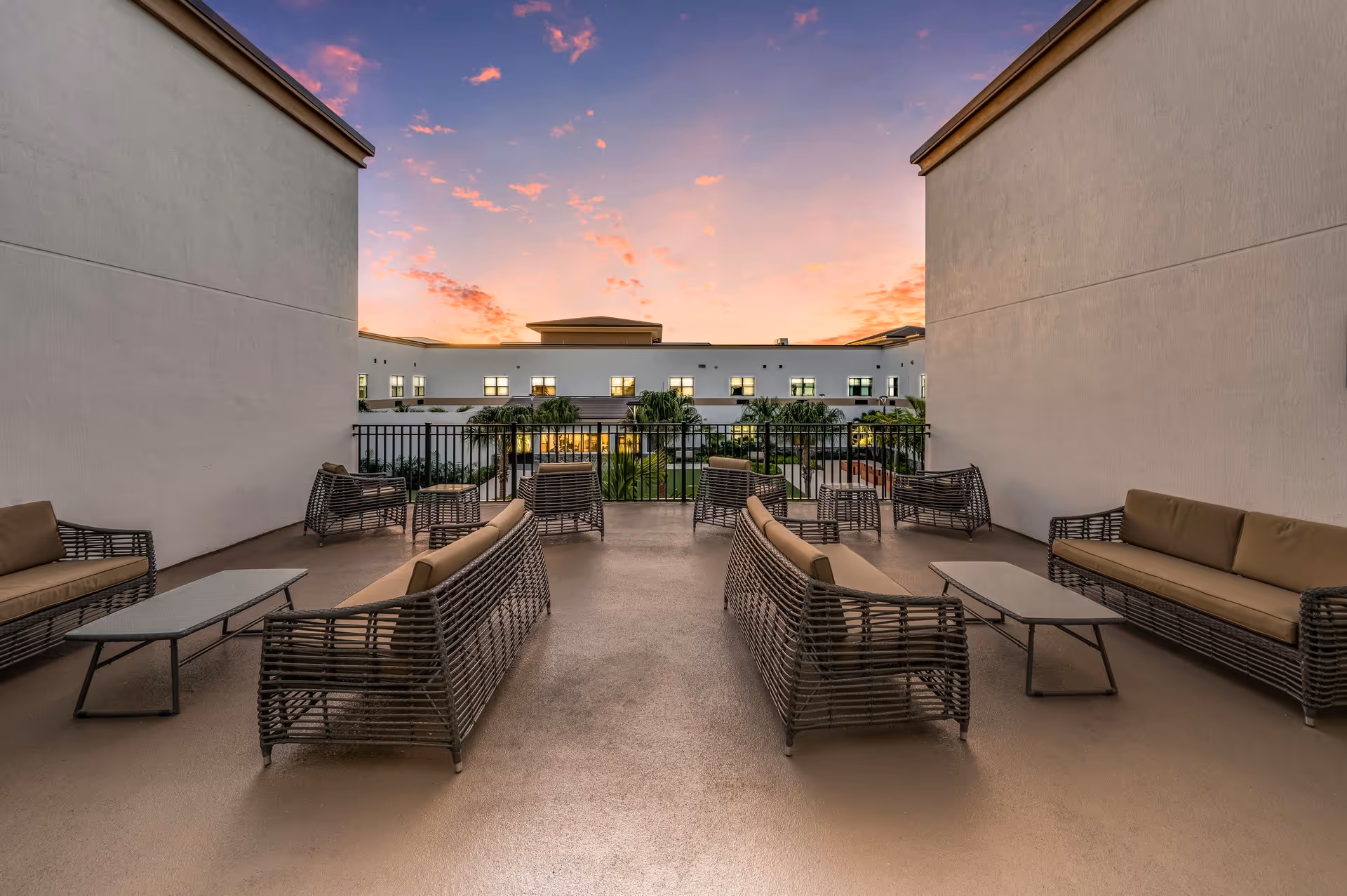 Outdoor patio area at sunset with multiple wicker sofas and chairs arranged around glass-top tables, surrounded by white walls and overlooking a garden with palm trees and a building in the background.