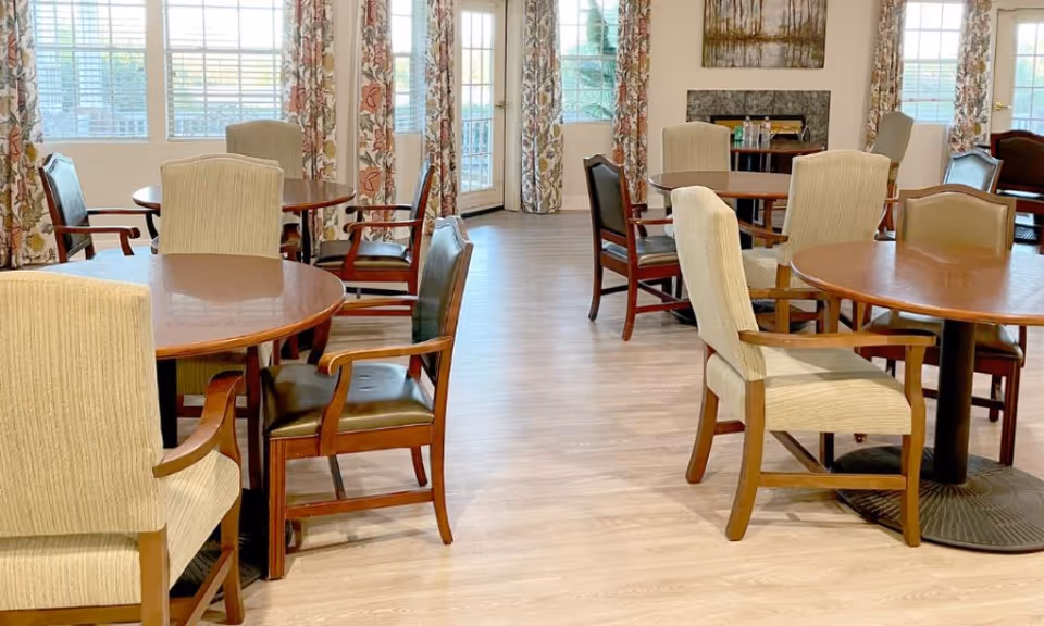 A bright dining room with round wooden tables and a mix of beige upholstered and dark wooden chairs. Large windows with floral curtains allow natural light to fill the room, and a painting hangs on the wall in the background.