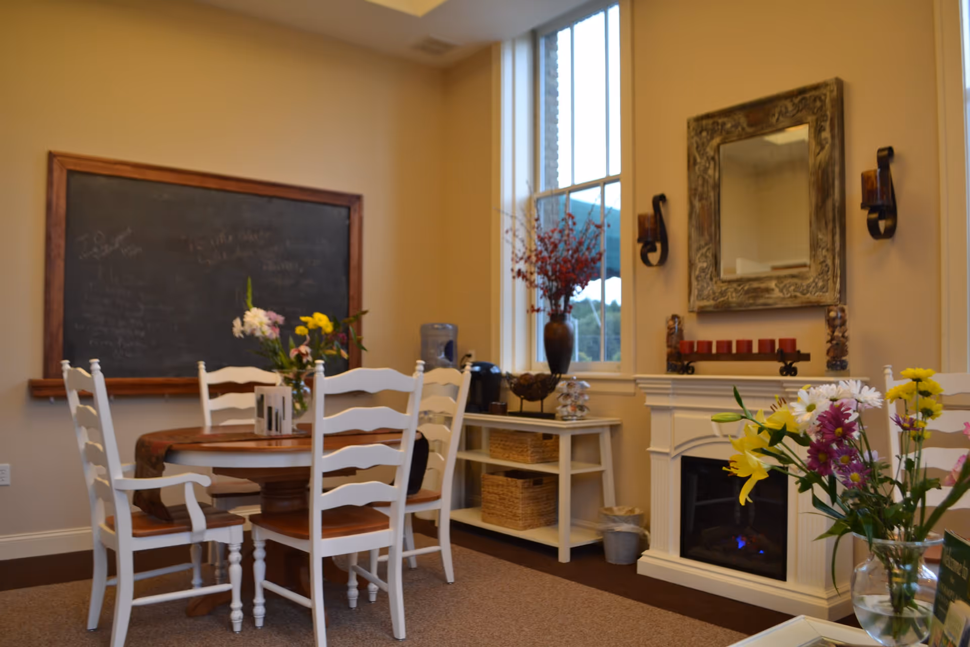 A cozy interior room with a round wooden table surrounded by white chairs. A vase with flowers is on the table. Behind the table is a large chalkboard with faint writing. To the right, there is a white fireplace with a decorative mirror above it and candle holders on either side. A side table with baskets and a water dispenser is next to a tall window with a vase of red flowers on top.