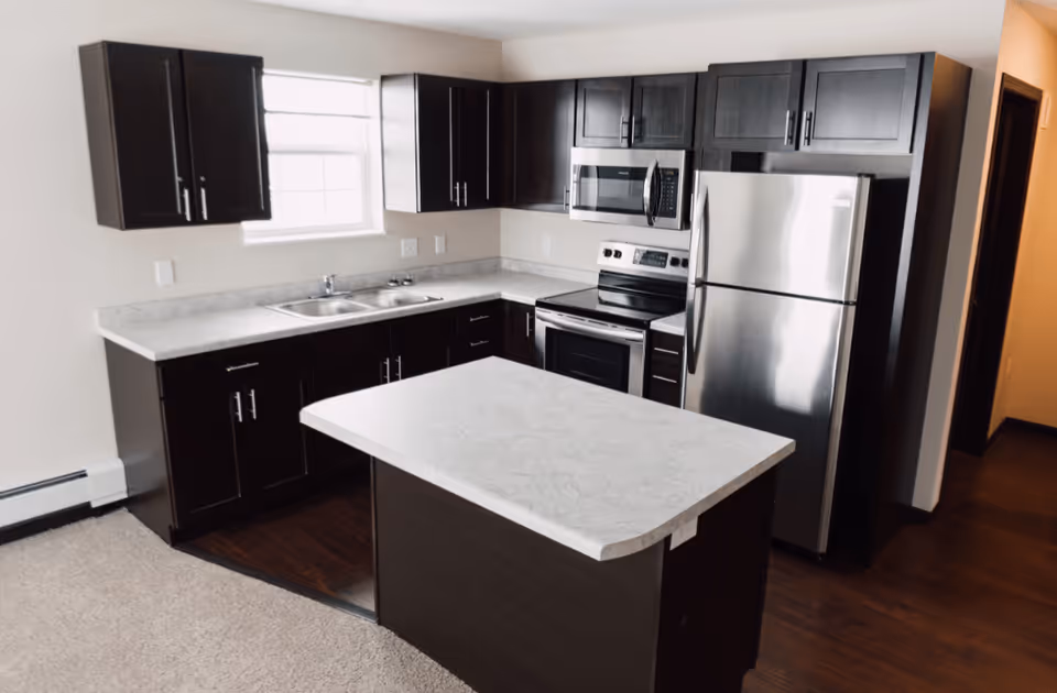 Modern kitchen with dark wood cabinets, a marble countertop island, stainless steel refrigerator, stove, and microwave, and a double sink under a window.