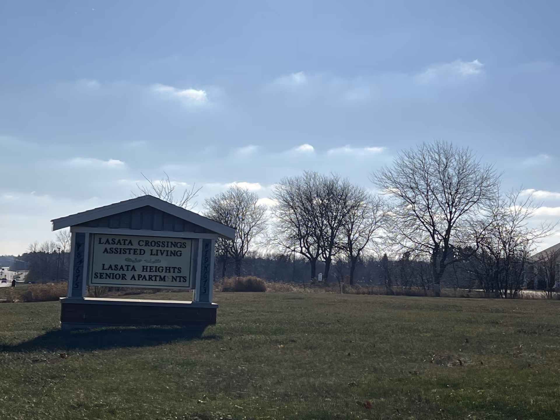 Outdoor view of a sign for Lasata Crossings Assisted Living and Lasata Heights Senior Apartments on a grassy area with leafless trees and a partly cloudy sky in the background.