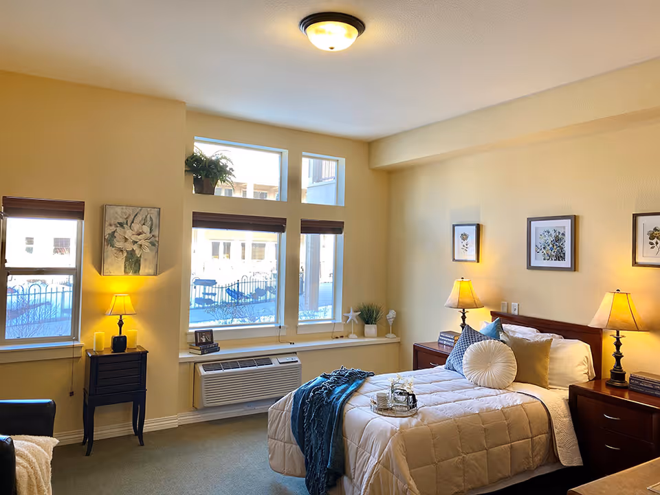 A cozy bedroom with a neatly made bed featuring multiple pillows and a blue throw blanket. The room has beige walls and carpeted floor. There are three windows with wooden blinds letting in natural light. Two bedside tables with lamps and decorative items flank the bed. Additional decorations include framed floral artwork on the walls and plants on the window ledge.