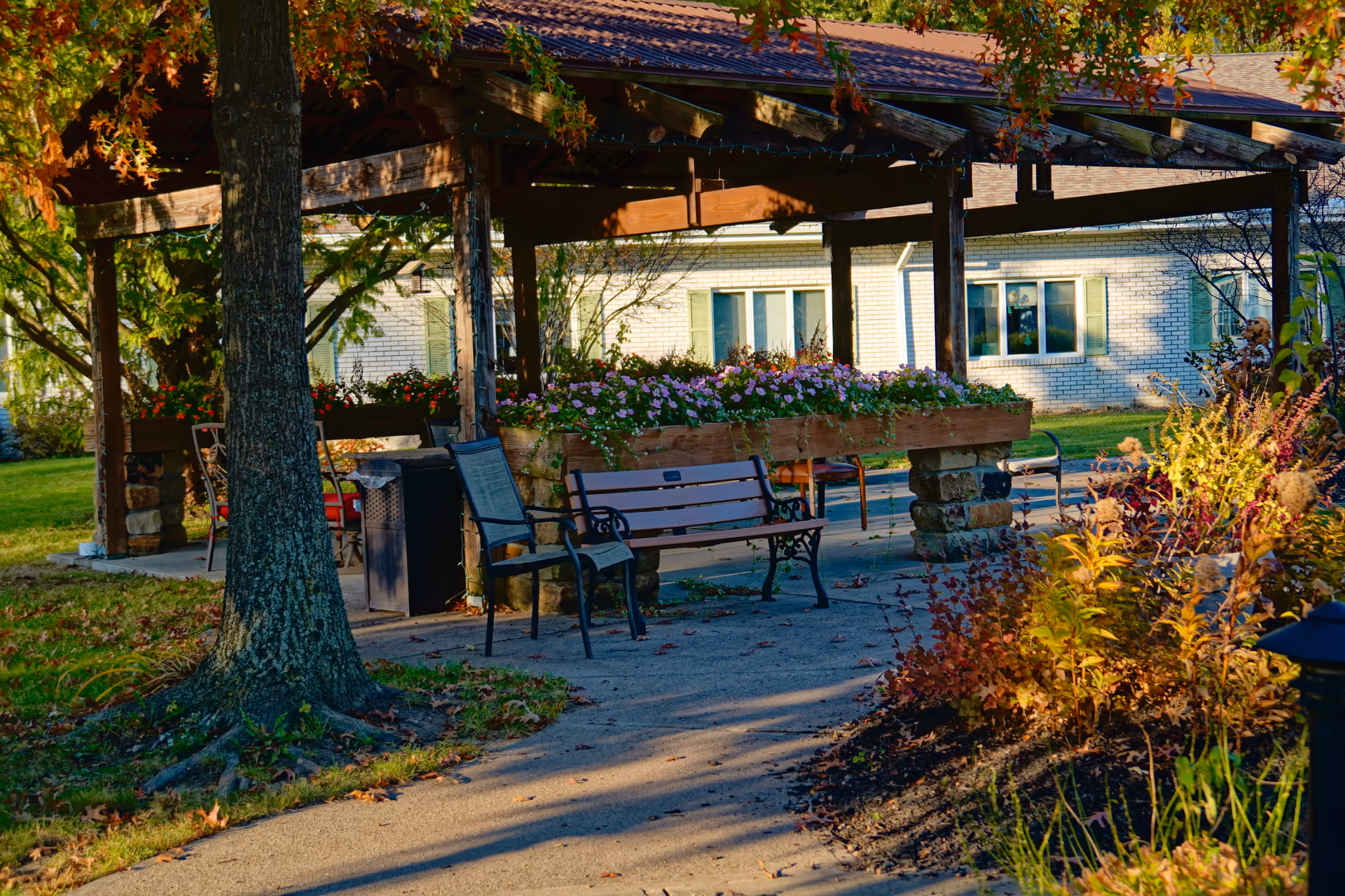 A shaded outdoor gazebo with benches, chairs, and flower planters along a paved path in front of a brick building.