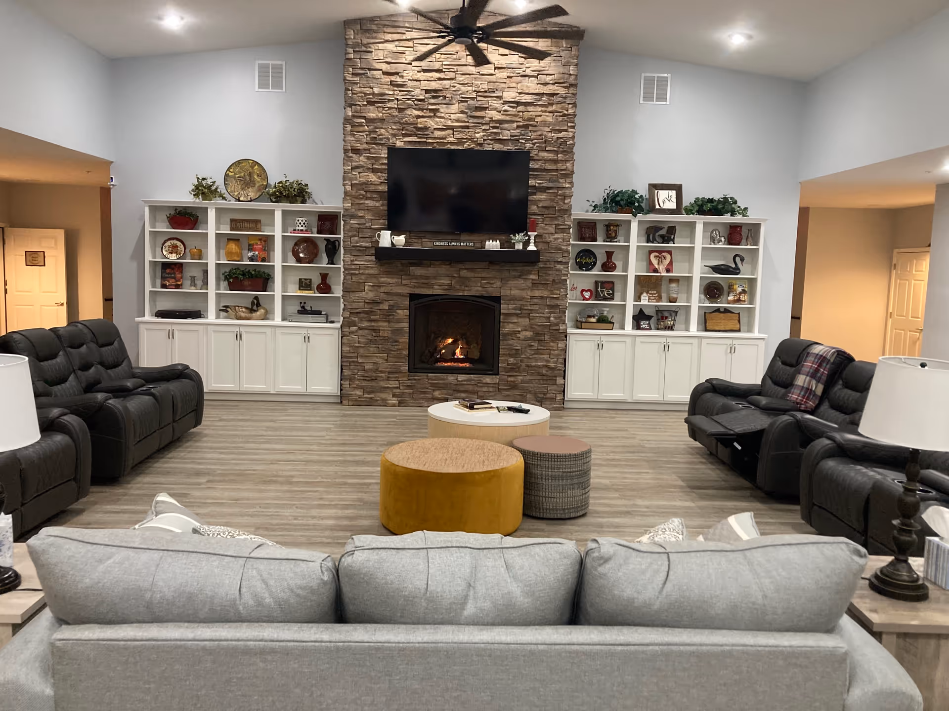 A spacious living room with a stone fireplace and a mounted flat-screen TV above it. The room features two white built-in shelves on either side of the fireplace, decorated with various vases, plants, and decorative items. There are multiple black leather recliners and a gray sofa facing the fireplace. In the center, there are three round ottomans in different colors and a small white coffee table. The room has light wood flooring and soft lighting from ceiling lights and table lamps.