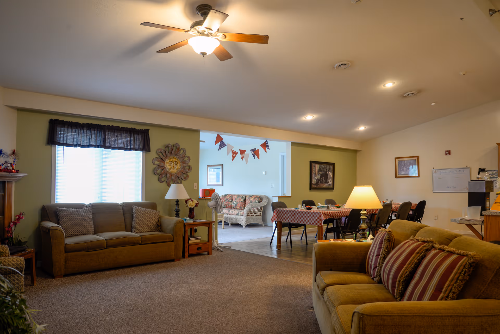 A cozy living room area with two sofas, one brown and one yellow with striped cushions, a side table with a lamp, and a ceiling fan with light. In the background, there is a dining area with a table covered with a red and white checkered tablecloth and several chairs. The walls are decorated with framed pictures and a sun-shaped wall ornament. The room is warmly lit with ceiling lights and a table lamp.