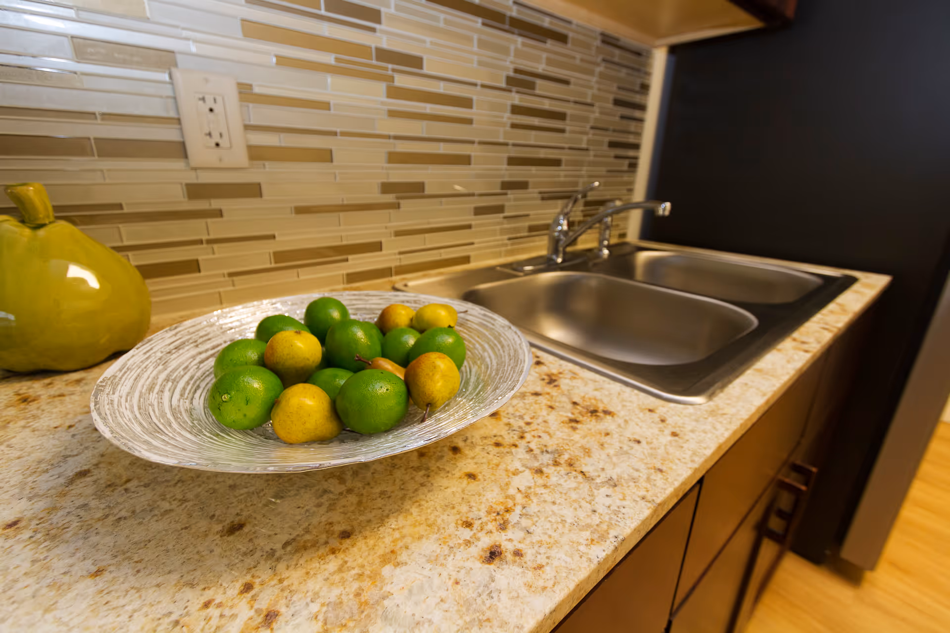 Close-up view of a kitchen countertop with a double stainless steel sink, a decorative plate filled with green and yellow fruits, and a green ceramic pear. The backsplash features horizontal rectangular tiles in shades of beige and brown.