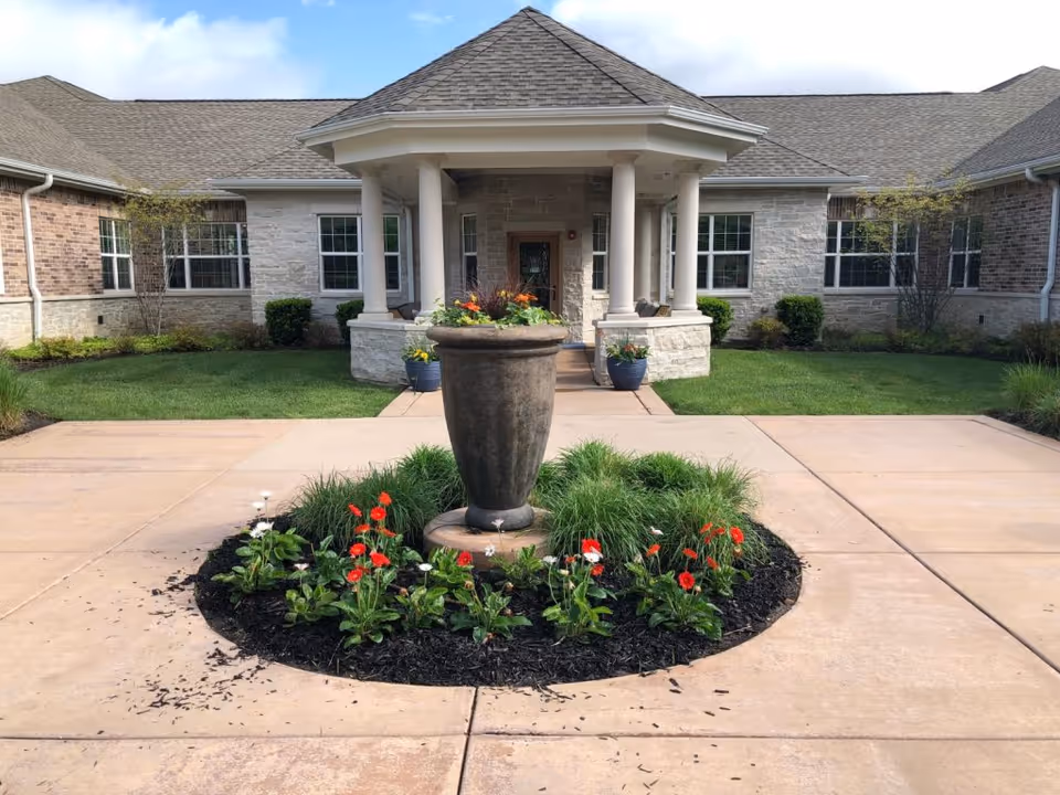Front entrance of Eagles Nest Assisted Living facility featuring a covered porch with four white columns, a large decorative planter with flowers and greenery in the center of a circular flower bed, surrounded by a paved driveway and well-maintained lawn and shrubs.