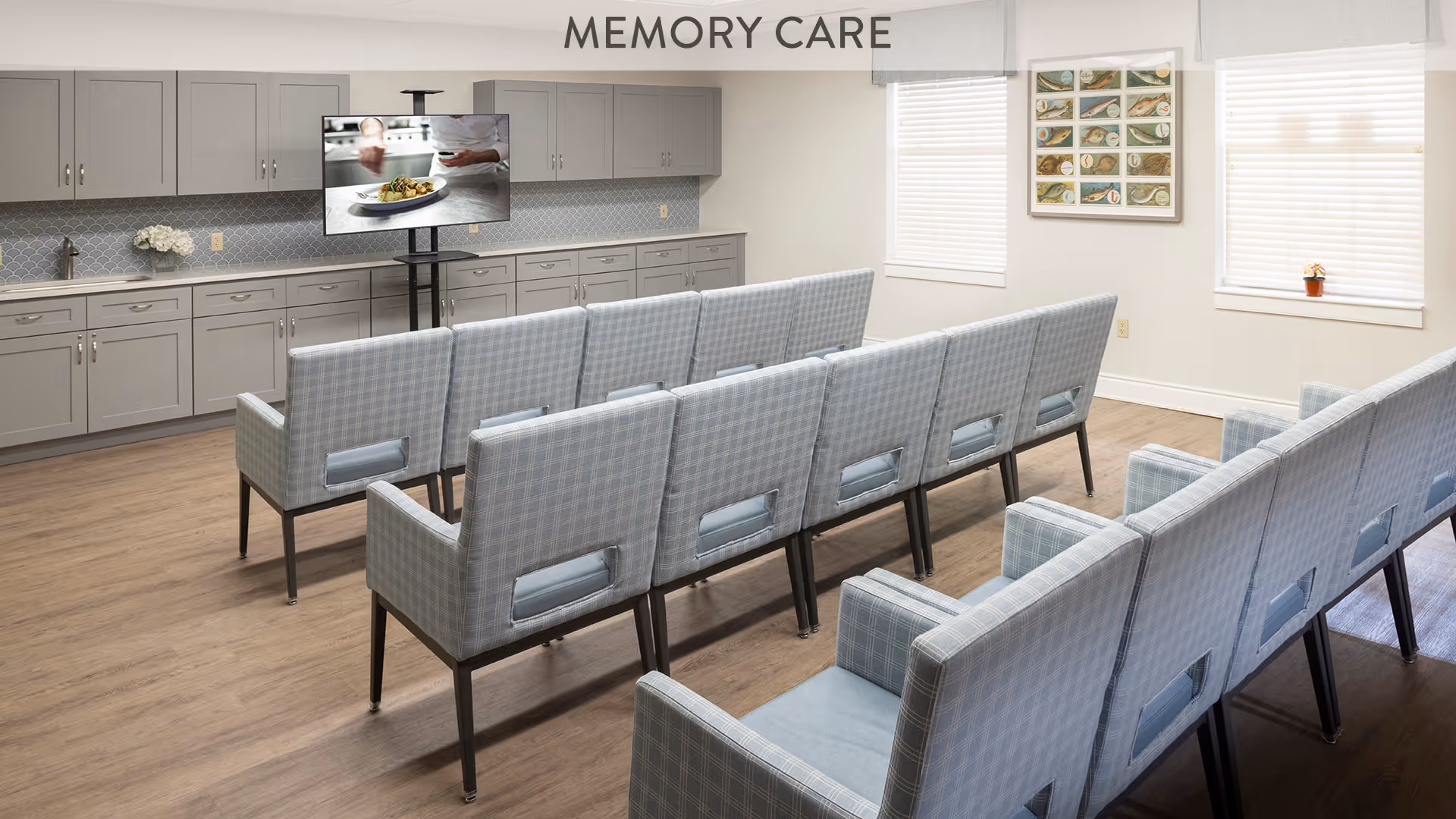 A memory care room with two rows of light blue plaid chairs facing a television mounted on a stand. The room has light wood flooring, gray cabinets along the back wall, two windows with blinds, and a framed picture on the wall.