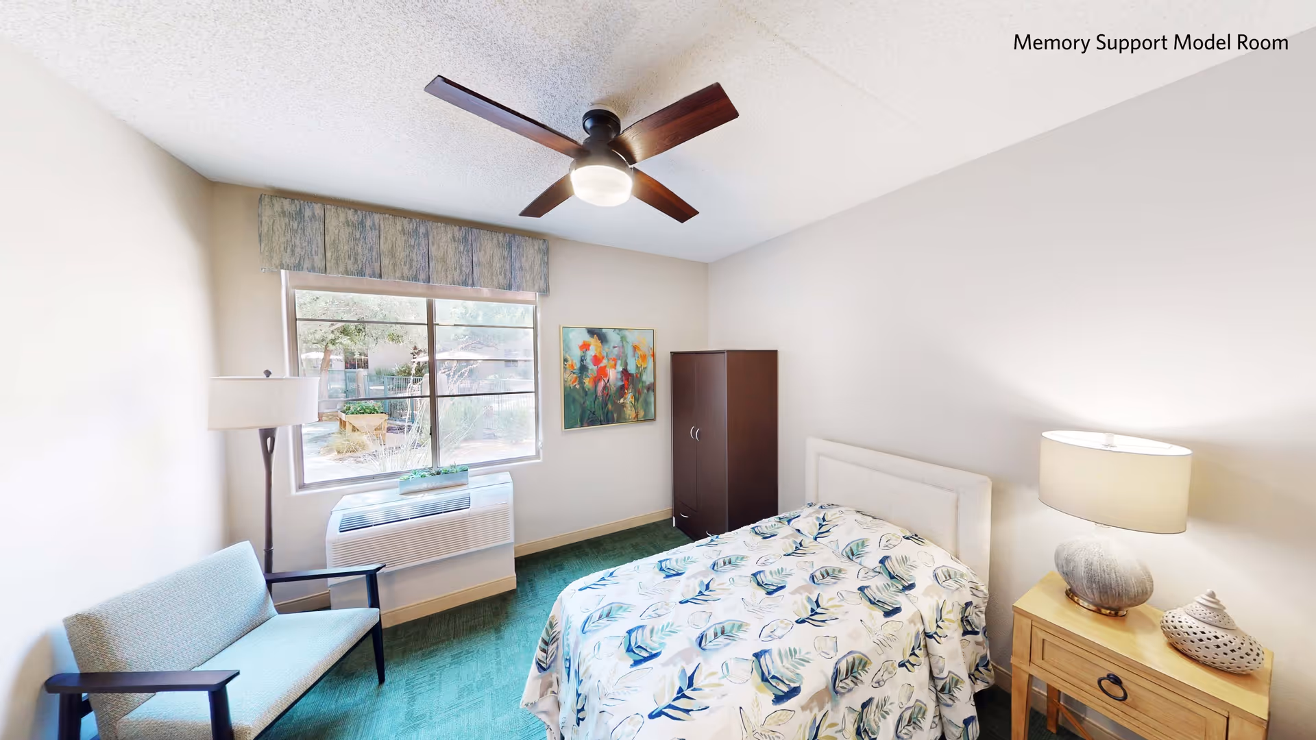 Model bedroom with a single bed, nightstand, armchair, ceiling fan and a window overlooking landscaping.