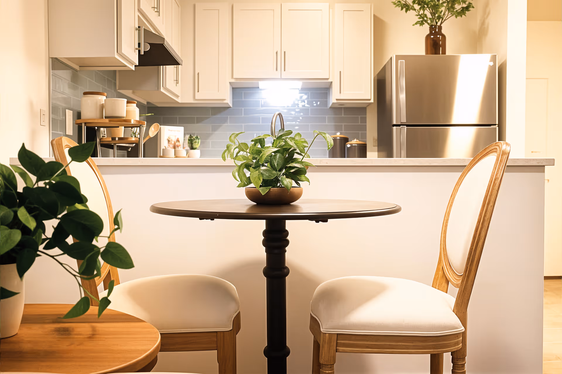 A cozy dining area with a round wooden table and two cushioned wooden chairs. A green potted plant is placed on the table. Behind the dining area is a kitchen with white cabinets, a gray tiled backsplash, a stainless steel refrigerator, and various kitchen items on the counter including jars and a small plant.