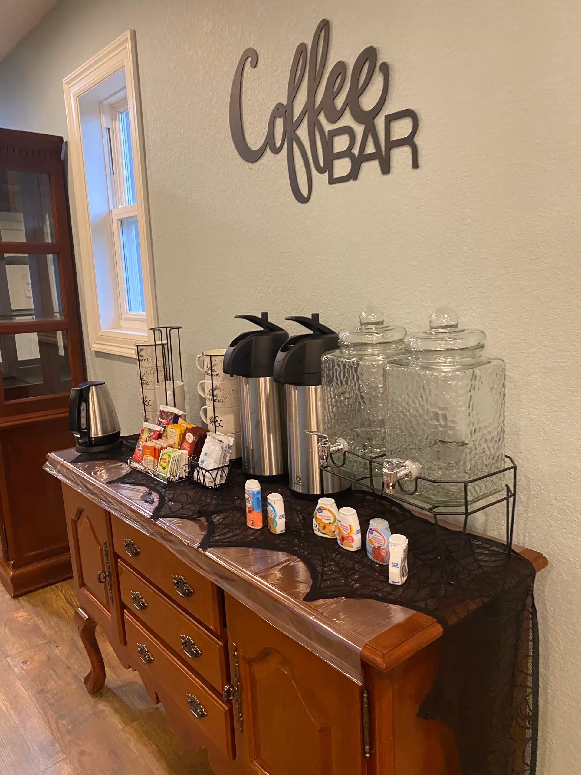 A coffee bar setup on a wooden sideboard with two large glass beverage dispensers, two stainless steel airpots, a kettle, various coffee and tea supplies, and creamers. The wall above has a decorative sign that reads 'Coffee BAR'.