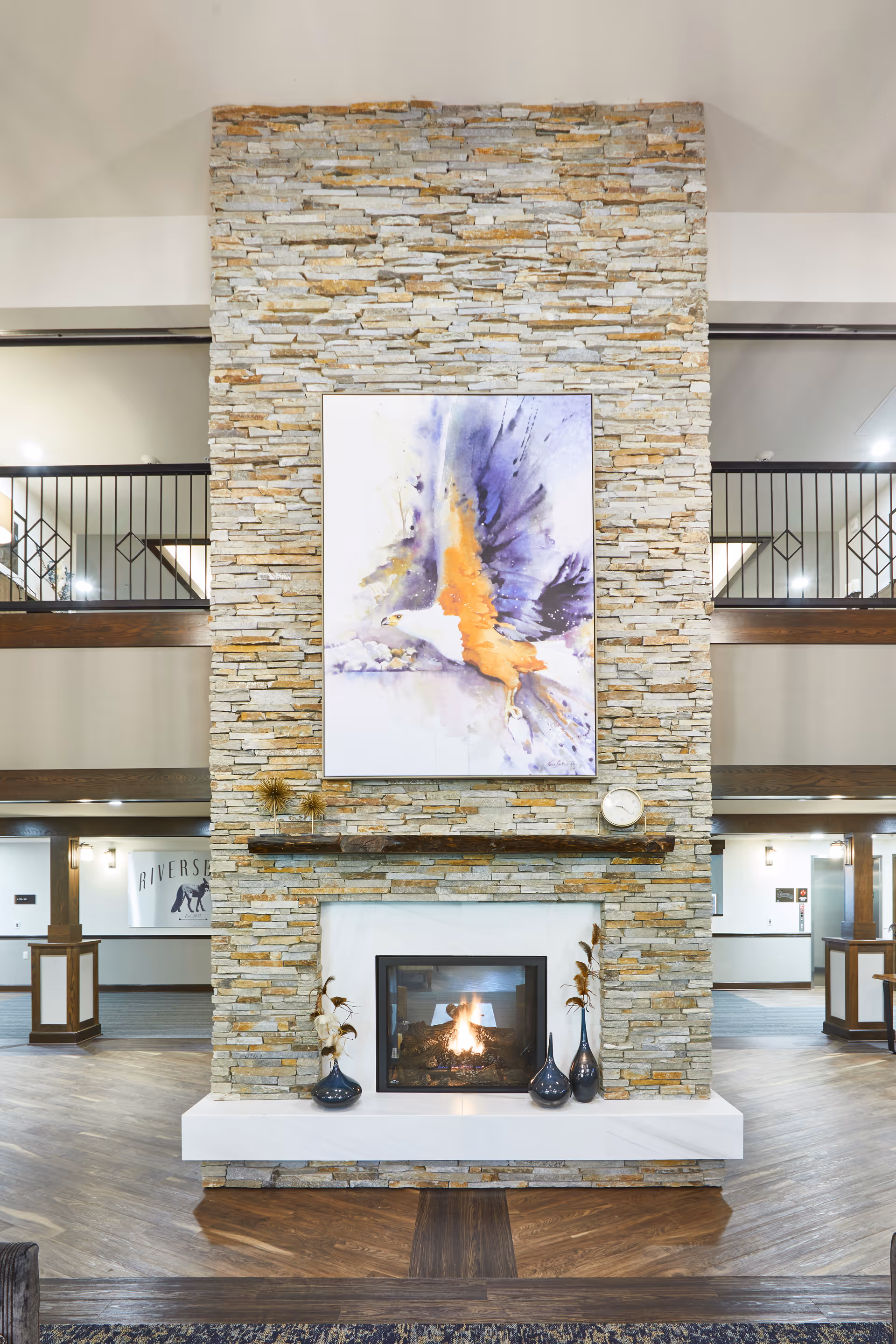 A tall stone fireplace with a lit fire in the center, decorated with three dark vases with dried plants on the hearth. Above the fireplace is a large colorful painting of an eagle in flight. The room has wooden floors and a two-story open space with railings on the upper level.
