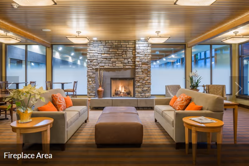 Cozy lounge with two sofas, a central ottoman, and a stone fireplace in a senior living facility.