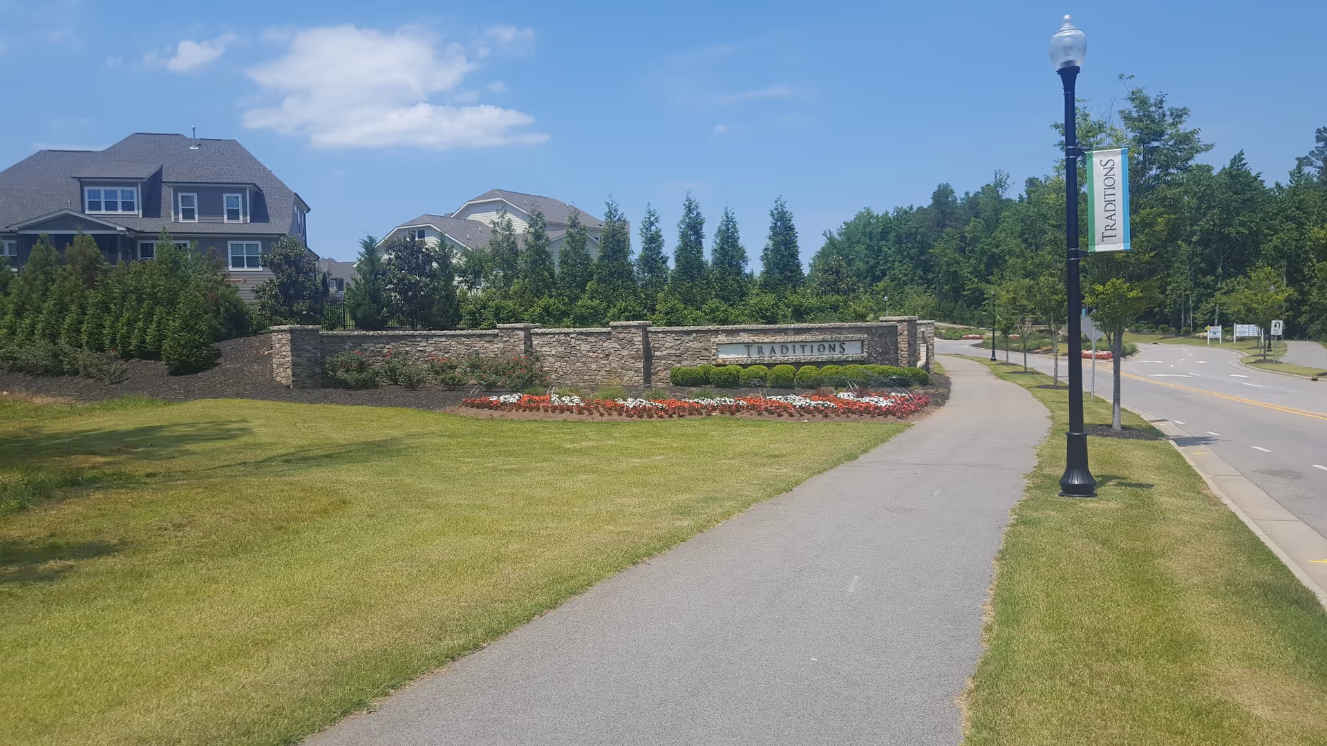 Stone entrance sign reading 'TRADITIONS' with landscaped flowerbeds, a sidewalk and lamp post banner leading to residential buildings under a blue sky.