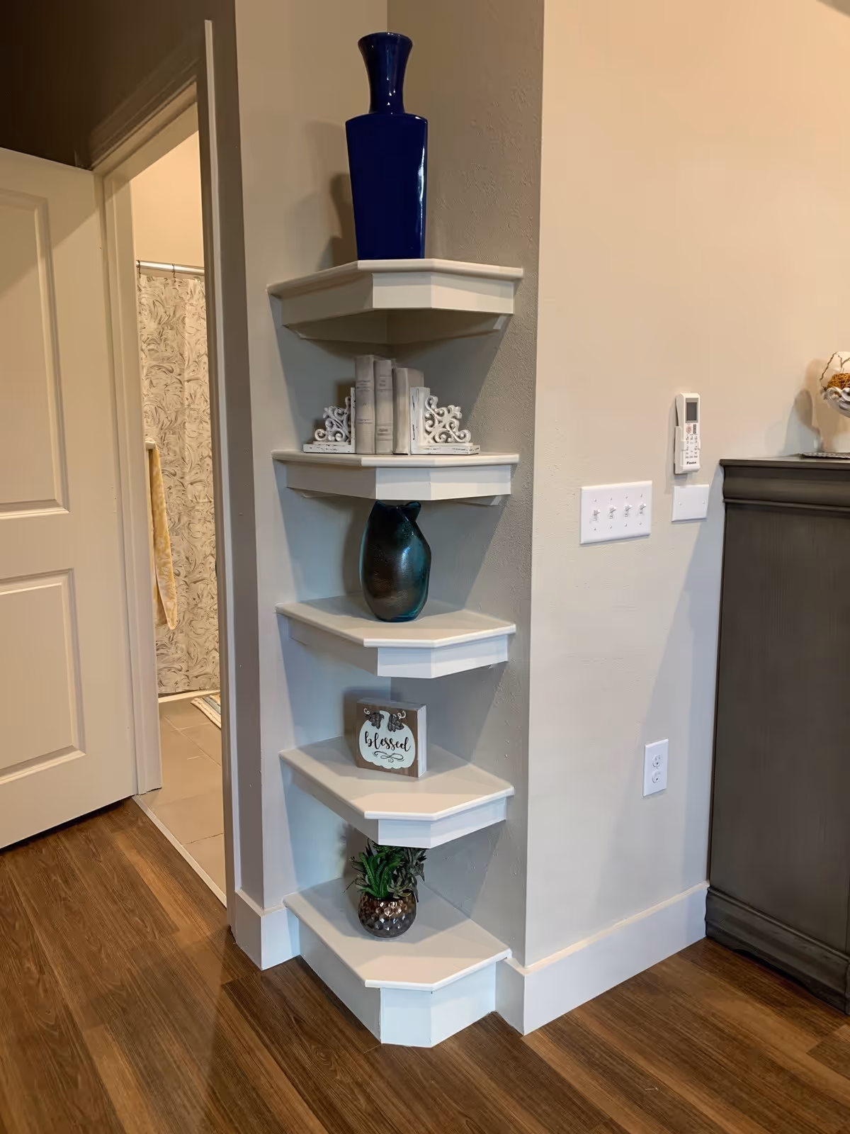 A corner of an interior room with five white corner shelves displaying decorative items including vases, books, a 'blessed' sign, and a small plant next to a doorway and cabinet.