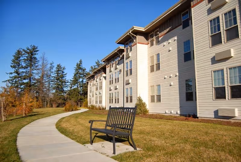 Outdoor view of a senior living facility building with multiple windows, a curved concrete walkway, a black metal bench on the grass, and trees in the background under a clear blue sky.