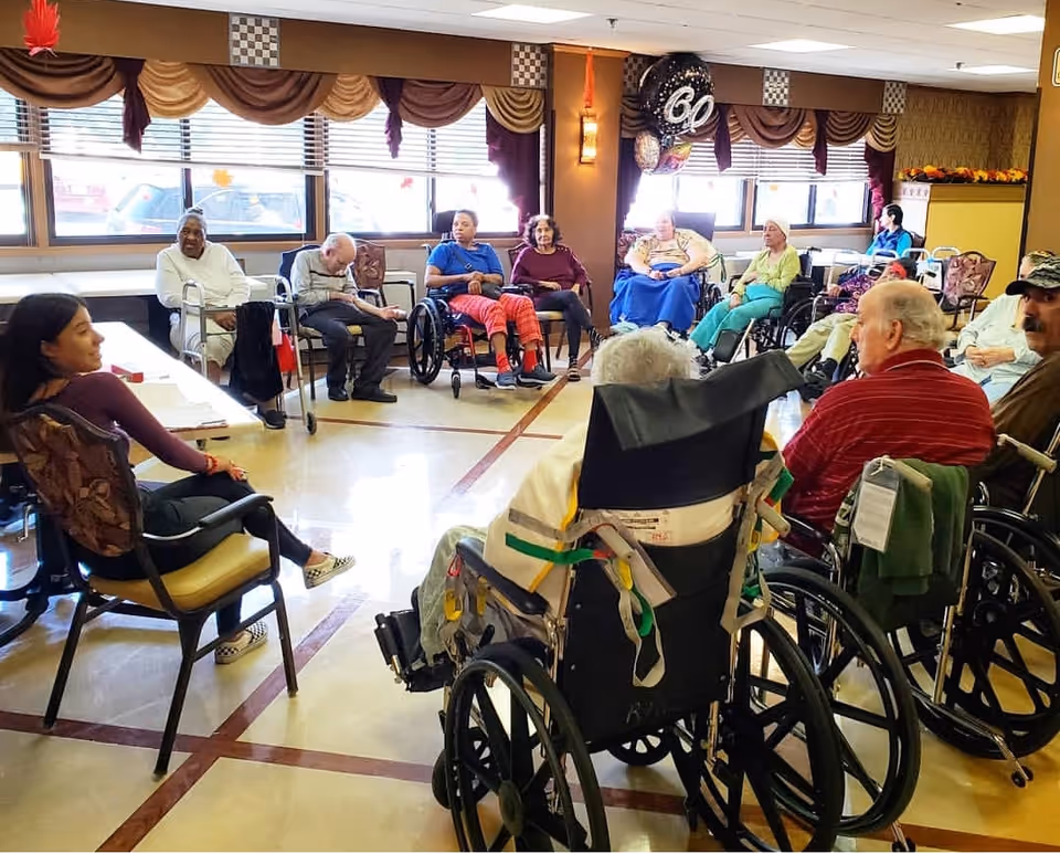 A group of elderly residents and staff seated in a circle in a bright activity/common room, many in wheelchairs.
