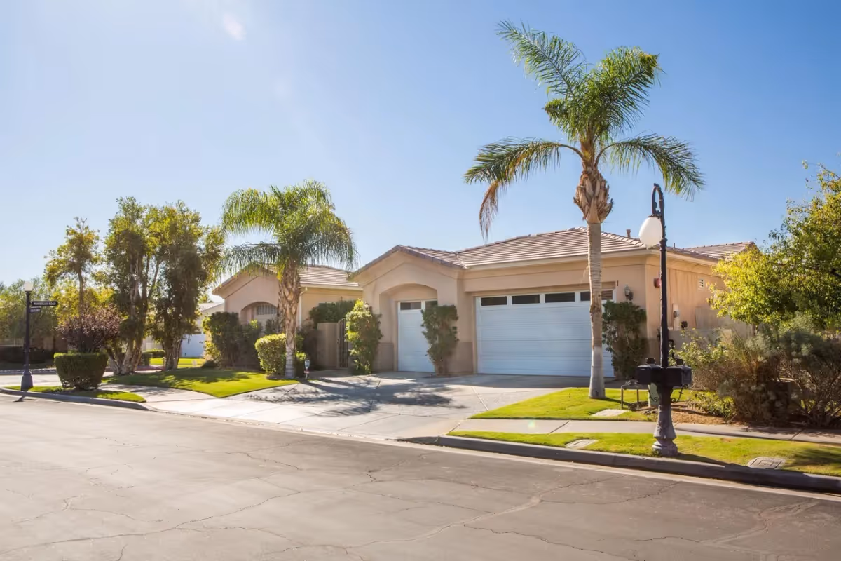 Exterior view of a single-story residential building with a tiled roof, two white garage doors, and a front yard with palm trees and bushes under a clear blue sky.